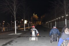 Vier rollifahrende Personen fahren im Dunkeln auf das Brandenburger Tor zu
