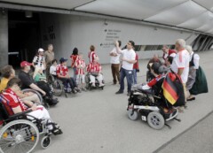 Ein Mann zeigt auf die Wand der Allianz-Arena, um ihn herum eine Gruppe Rollstuhlfahrer*innen
