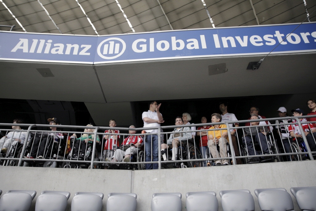 Gruppe Rollstuhlfahrer*innen auf den Rolliplätzen, ein Mann steht in der Mitte, darüber ein Werbebanner: Allianz Global Investors