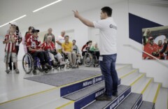Ein Mann zeigt auf etwas und steht auf Treppen mit Aufschrift Allianz Arena, vor ihm eine Gruppe Rollstuhlfahrer*innen