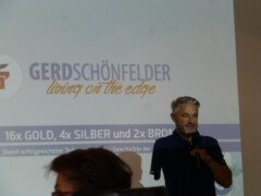 Gerd Schönfelder steht mit einem Mikrofon vor der Leinwand auf der steht Gerd Schönefelder und darunter living on the edge