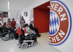 Einige Rollstuhlfahrer*innen fahren auf einen Durchgang zu auf dessen Seiten die beiden Hälften des FC Bayern Logos