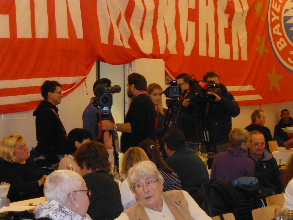 Einige Personen sitzen an Tischen, mehrere Personen mit Kameras im Hintergrund