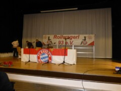 Drei Personen auf der Bühne, ein langer Tisch mit Mikrofonen mit FC-Bayern Logo geschmückt