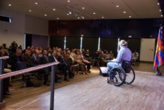 Kim Krämer sitzt im Rollstuhl auf einer Bühne in einem Saal vor Publikum