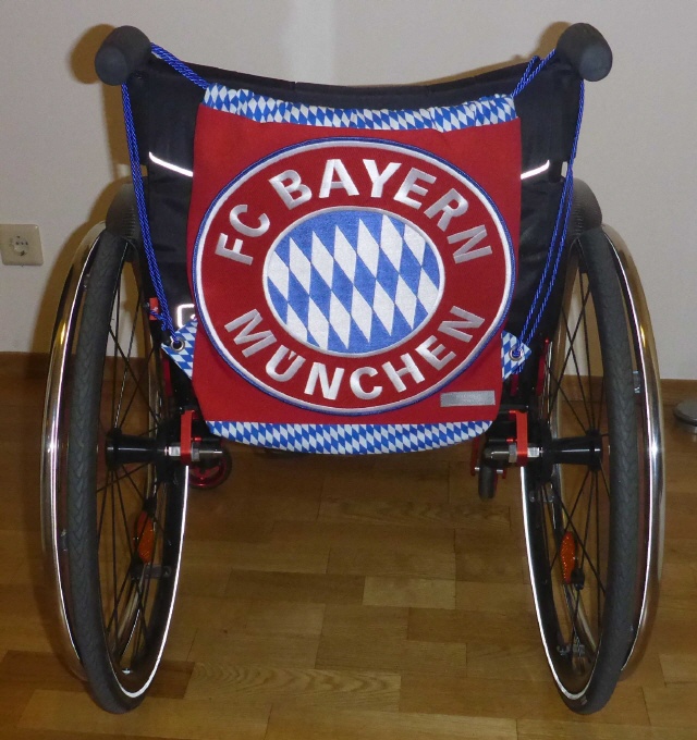 Rollstuhl von hinten mit FC Bayern Rucksack aufgehängt an den Haltegriffen rechts und links oben
