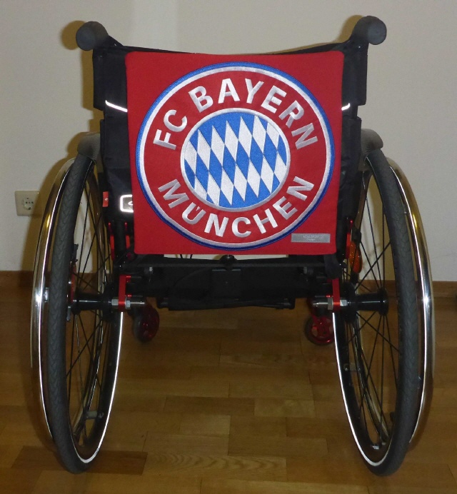 Rollstuhl von hinten mit rotem FC Bayern Rückenschild