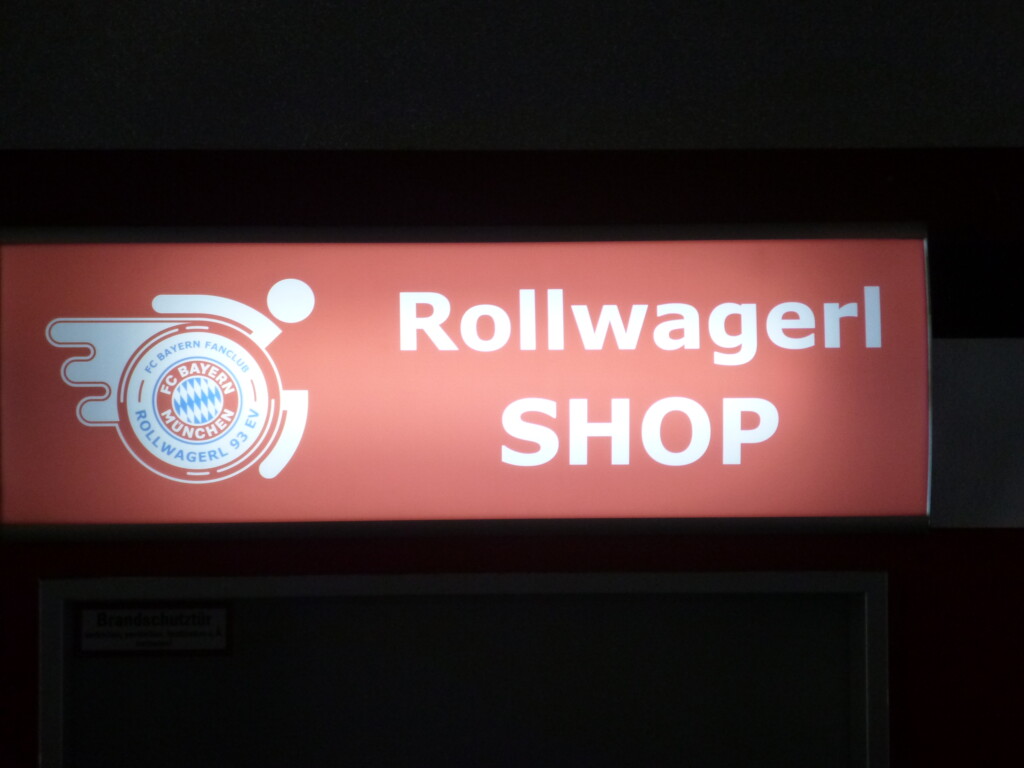 Rollwagerl-SHOP Schild über der Eingangstür des Rollwagerl-SHOP in der Allianz Arena beleuchtet bei Nacht.