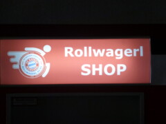 Rollwagerl-SHOP Schild über der Eingangstür des Rollwagerl-SHOP in der Allianz Arena beleuchtet bei Nacht.
