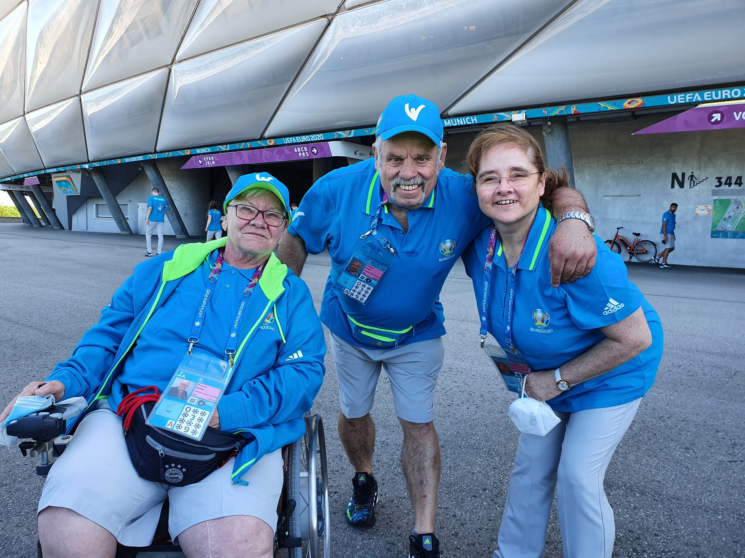 Drei Volunteers vor der Allianz Arena, eine Frau im Rollstuhl und zwei Fußgänger*innen