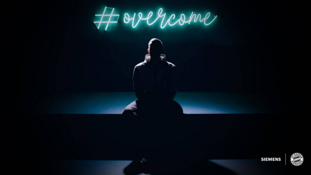 Person sitzt in einem abgedunkelten Raum und ist nicht zu erkennen; Schrift zu #overcome; Logo von Siemens und Bayern München