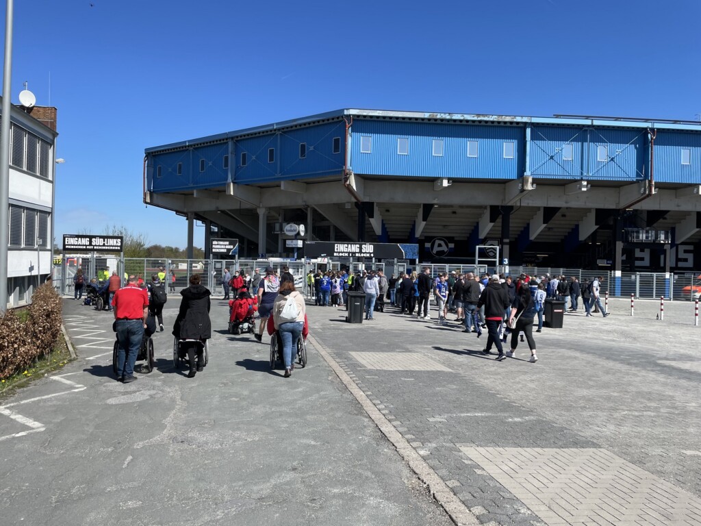 Im Hintergrund ein Fußballstadion. Im Vordergrund fußballfans auf dem Weg zum Eingang