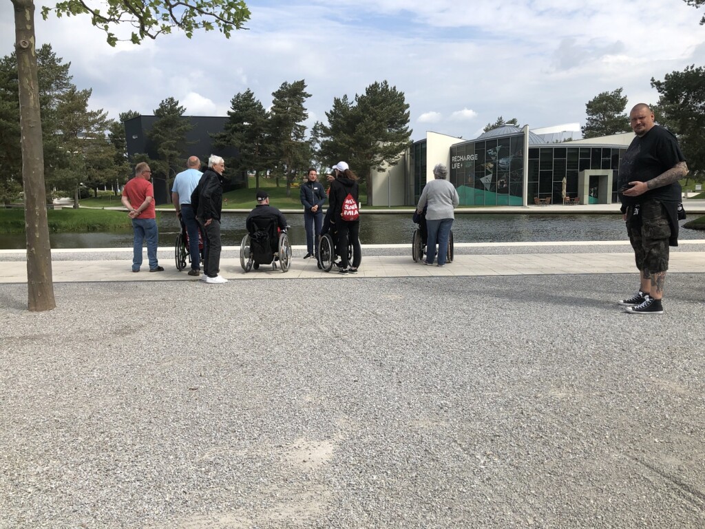 10 Personen vor einem See. Im Hinergrund sind zwei Gebäude und Tannen.