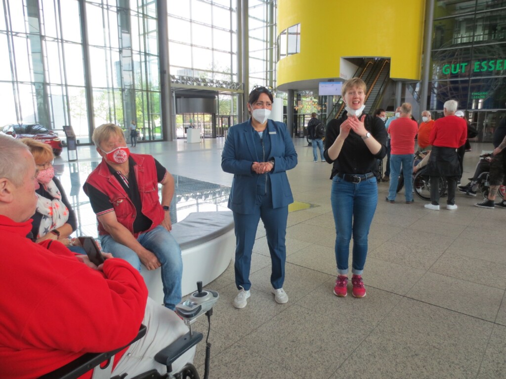 Eine Gebärdensprachdolmetscherin übersetzt bei einer Führung in Autostadt Wolfsburg. Zwei Rollstuhlfahrer und eine Frau stehen daneben.