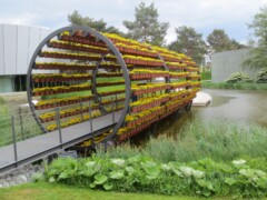 Eine futuristische Brücke in einem Park. Die Brücke führt durch Rohr, welches aus Blumentöpfen mit gelben Blumen besteht.