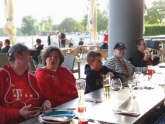 Menschen sitzen gesellig an einem Tisch im Außenbereich eines Restaurants. Zwei tragen ein FC Bayern Trikot.