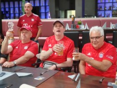 Vier FC Bayern Fans sitzen an einem Tisch und stoßen mit einem Getränk an.