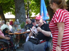 Ein Rollstuhlfahrer spricht in ein Mikrofon und hält eine Geburtstagskarte in der Hand. Daneben steht eine Frau.