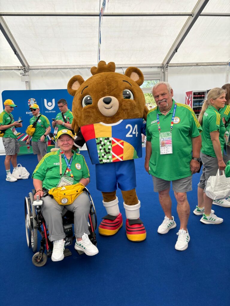 Drei Personen in einem großen Zelt. Eine davon eine Rollstuhlfahrerin und mittig das Maskottchen der EURO 2024