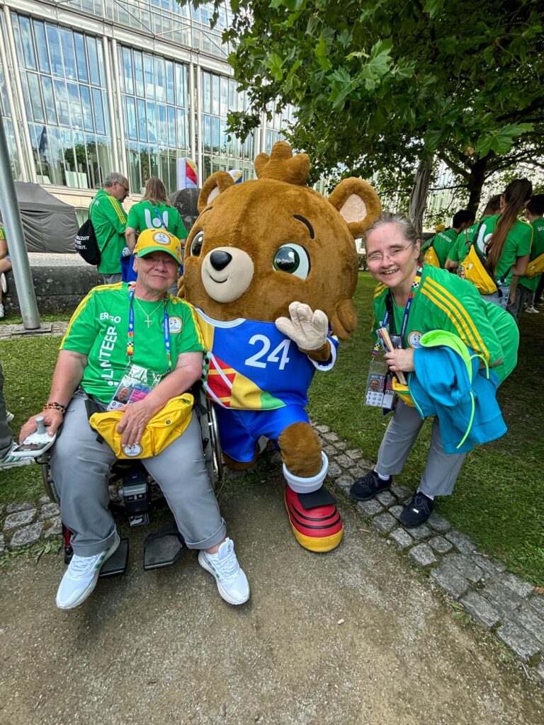 Drei Personen vor einem Baum. Eine davon eine Rollstuhlfahrerin und mittig das Maskottchen der EURO 2024