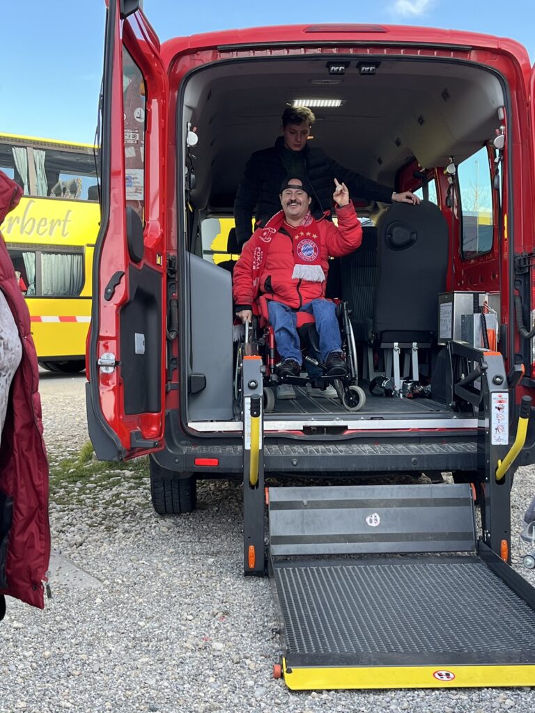 Eine Rollstuhlfahrer hebt die Hand in einem Kleinbus. Eine Rampe ist ausgefahren und die Hecktüre ist geöffnet.