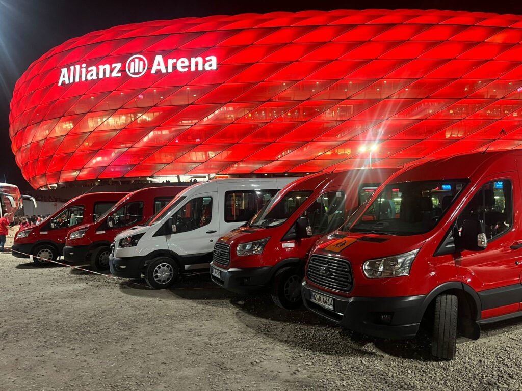 Vier Kleinbusse vor der Allianz Arena bei Nacht.