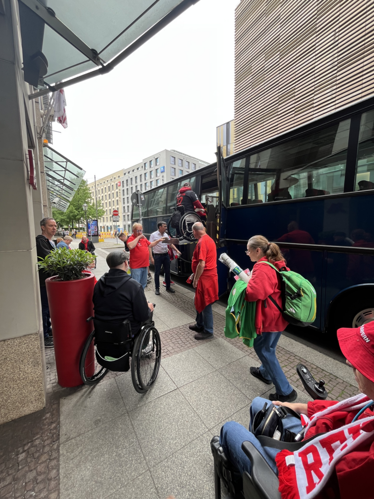 10 Personen davon drei mit Rollstuhl vor einem Reisebus. Ein Rollstuhlfahrer wird mit Lift in den Bus gebracht.