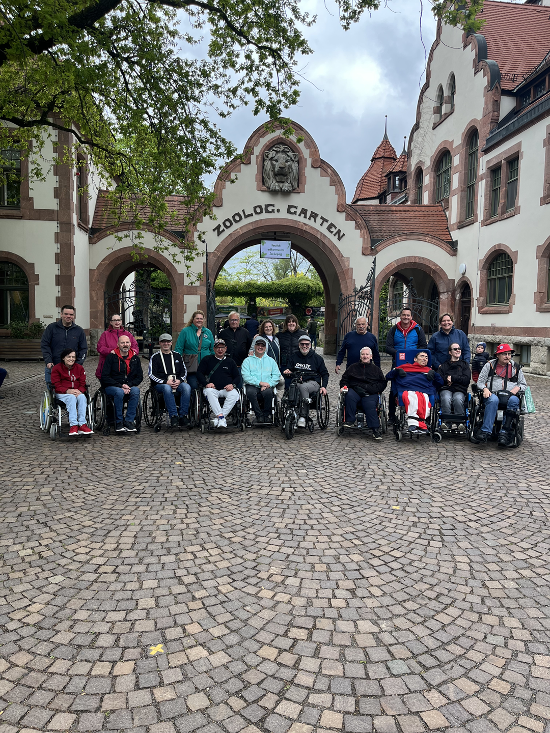 Ein Gruppenfoto mit 10 Rollstuhlfahrern und 9 Begleitern vor dem Eingang des Zoos von Leipzig