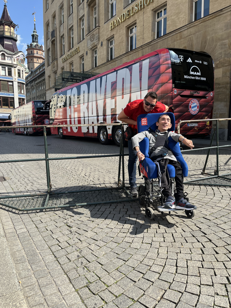 Ein Rollstuhlfahrer mit Begleitung vor einer Absperrung. Dahinter 2 Reisebusse des FC Bayern München.