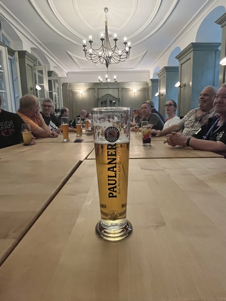 Ein großer Tisch mit einem vollem Weißbierglas im Vordergrund. Im Hintergrund Personen am Tisch, die lachen und sich unterhalten.