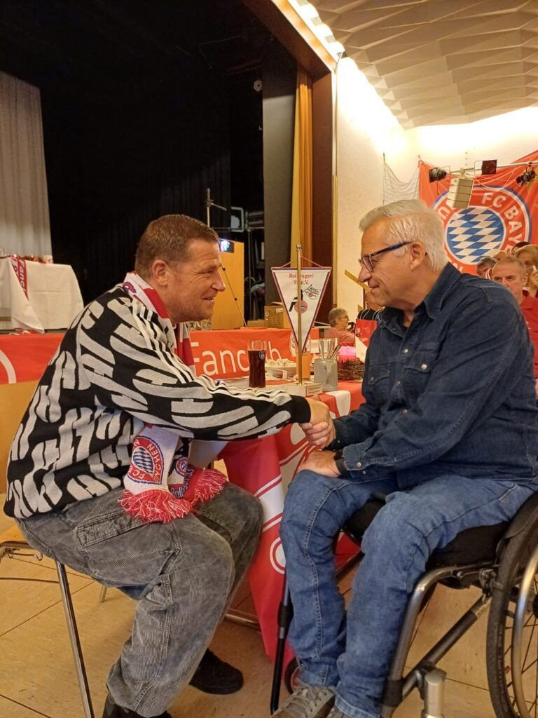 Ein Menschen mit Rollstuhl und Max Eberl sitzen an einem Tisch und unterhalten sich.