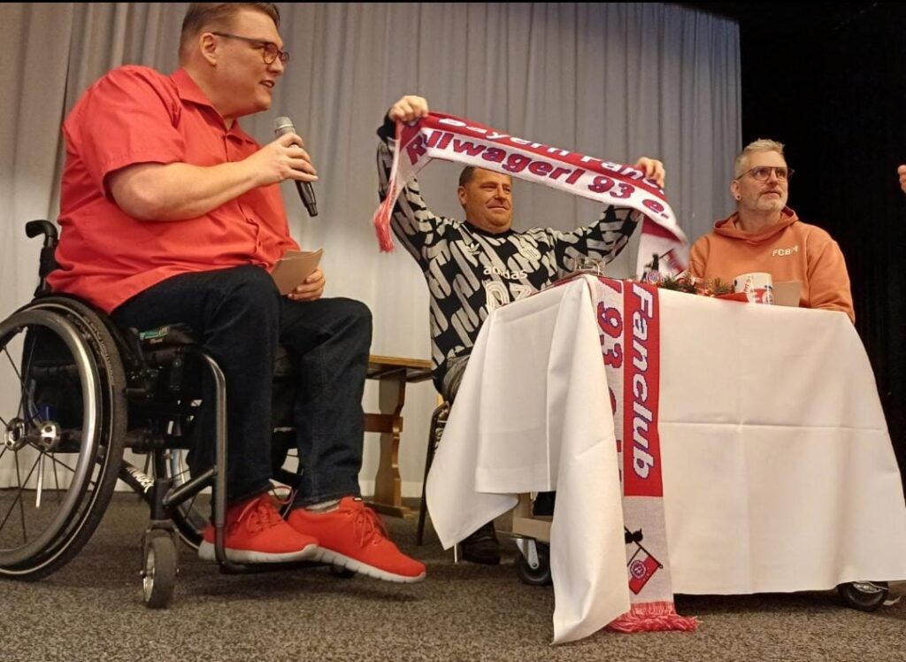 Max Eberl hält einen Fanschal nach oben. Darauf steht Rollwagerl 93 e.V. Daneben Kim Krämer und Alexander Grundler.