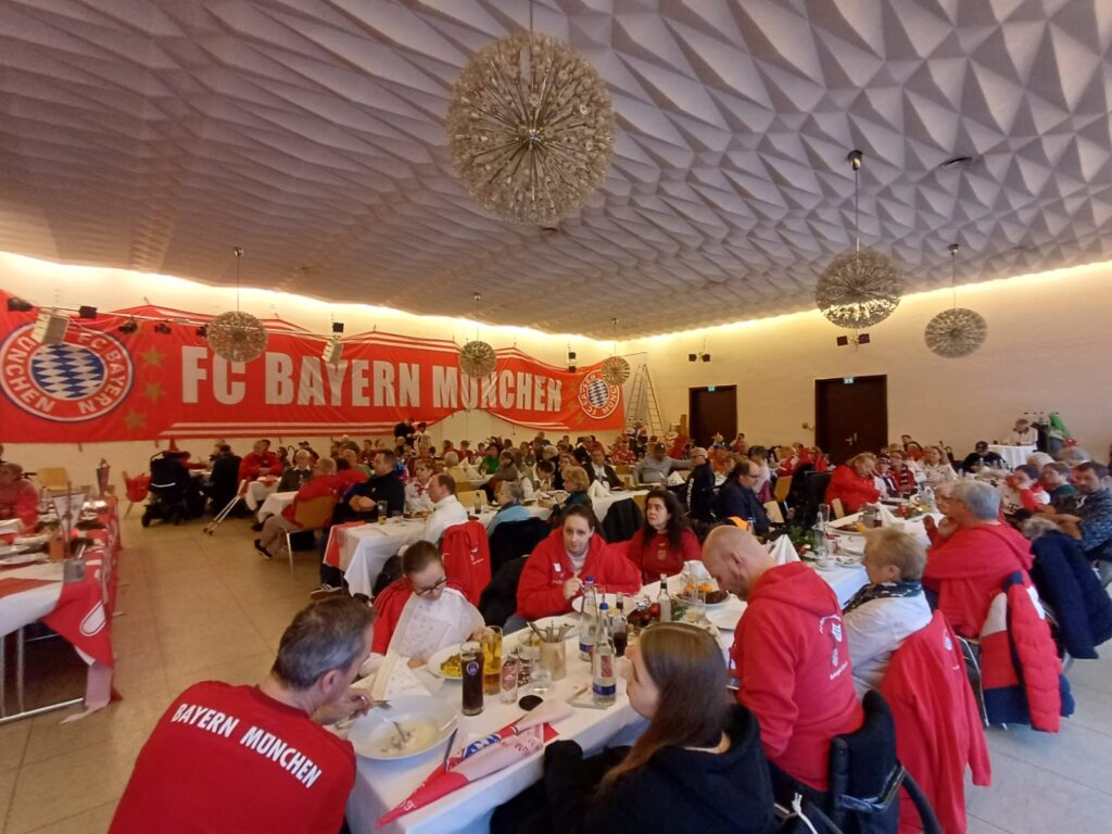 Weihnachtsfeier des Rollwagerl 93 e.V.! Die Teilnehmer sitzen in einem großen Saal. Im Hintergrund ist ein großer FC Bayern Banner zu sehen.
